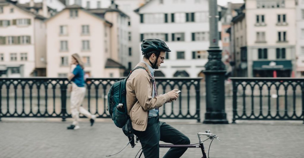 A man on a bicycle uses his smartphone while paused on a city bridge during the day.
