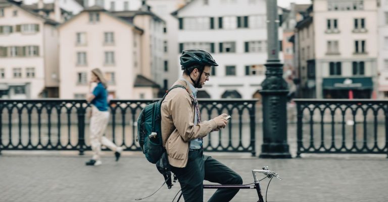 A man on a bicycle uses his smartphone while paused on a city bridge during the day.