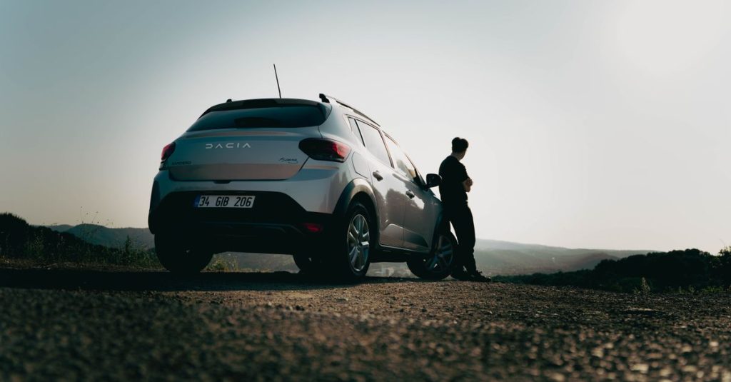 A man leans against a Dacia car on a scenic road at sunset, capturing a sense of wanderlust.