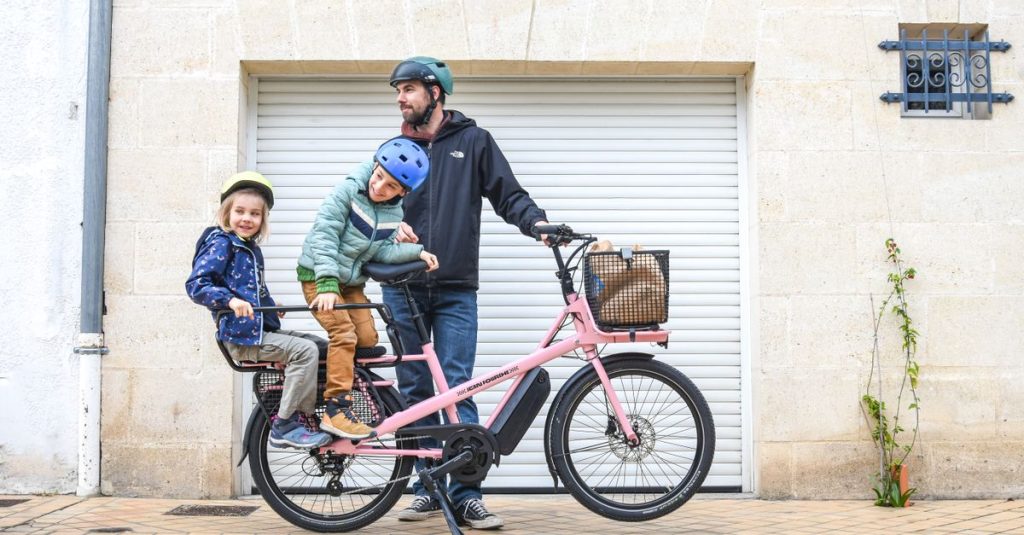 A father and two children wearing helmets ride a pink cargo bike in Paris.