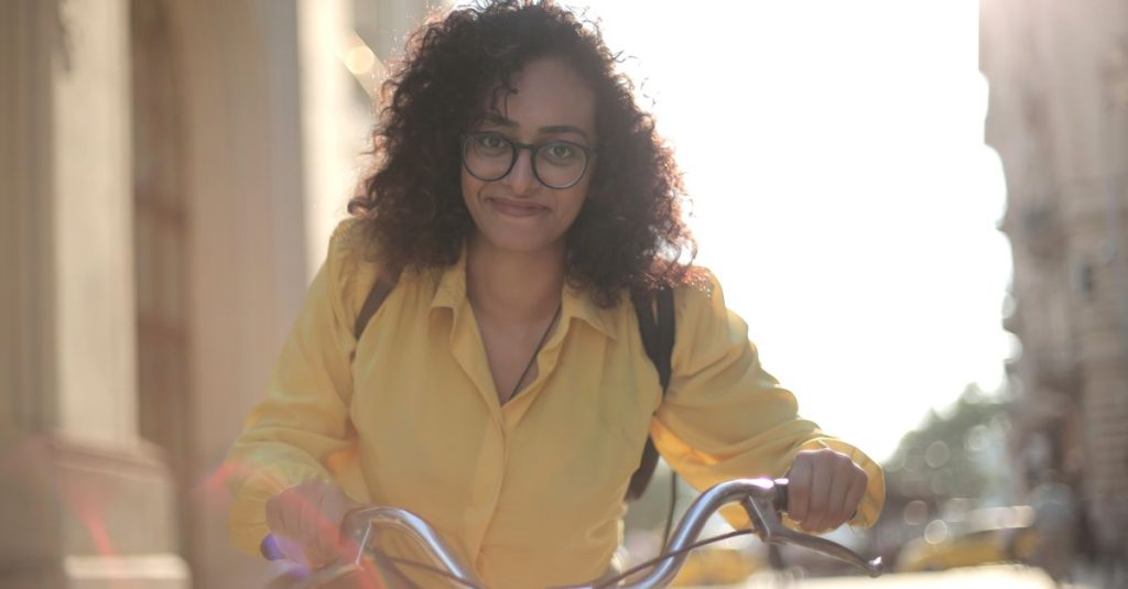 From below cheerful young curly woman in glasses with backpack smiling at camera while riding bicycle along street in sunny day