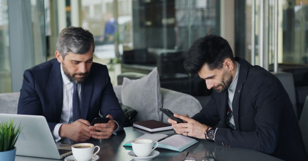 Two businessmen in suits using smartphones at a café table with laptops and coffee.
