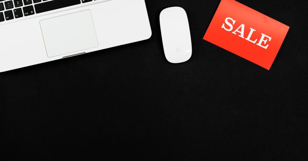 Minimalist flat lay of a laptop, mouse, and a red sale sign on black background.