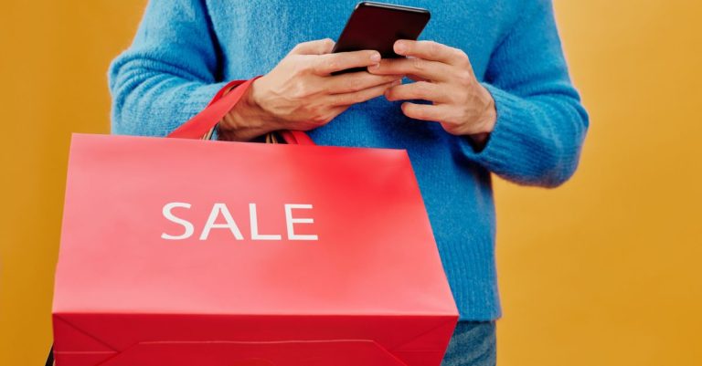 Close-up of a man holding a sale bag and smartphone, symbolizing shopping and discounts.