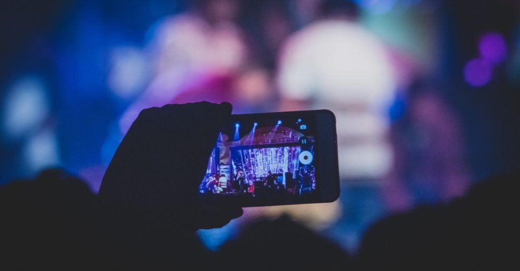 A hand holds a smartphone recording a live concert with vibrant lights.