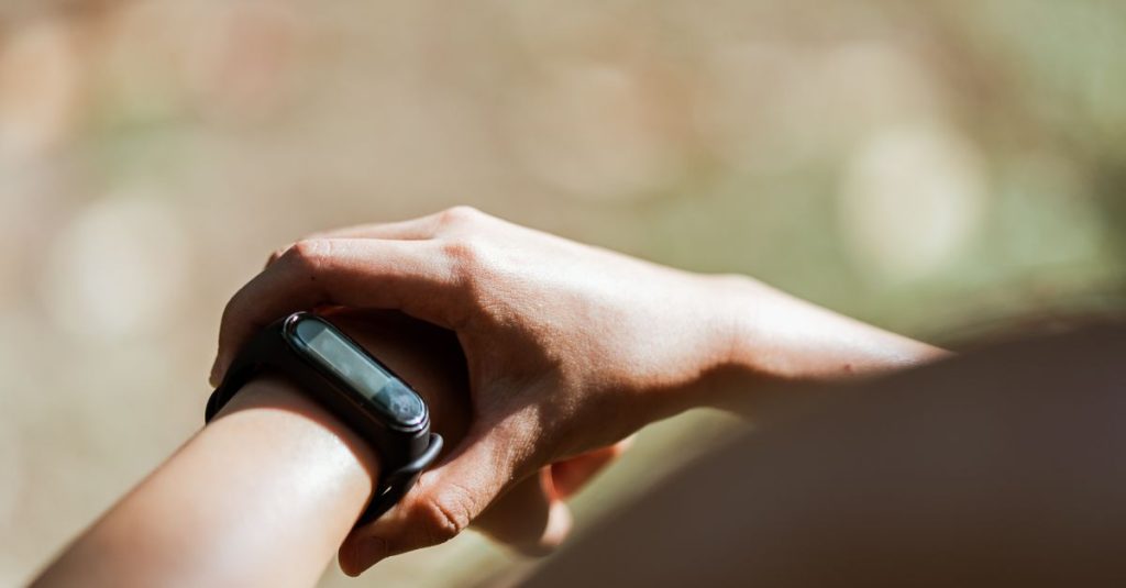 From above of crop anonymous person wearing modern bracelet with small display while standing outdoors on blurred background in daylight
