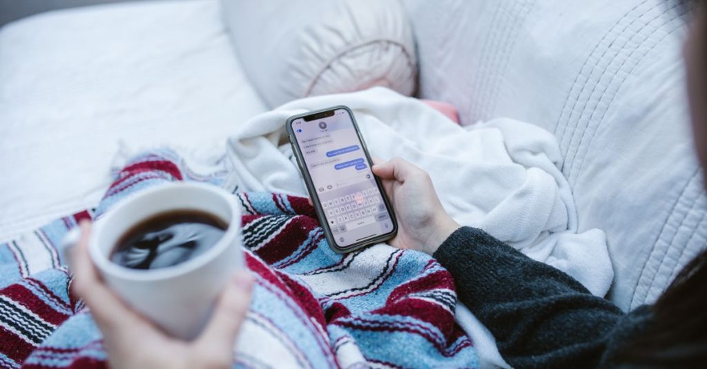 Person relaxing indoors with coffee and smartphone, texting in a cozy setting.