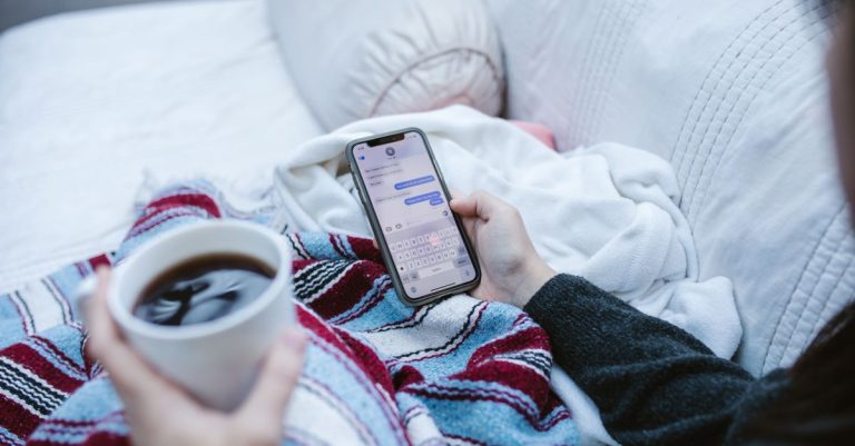 Person relaxing indoors with coffee and smartphone, texting in a cozy setting.