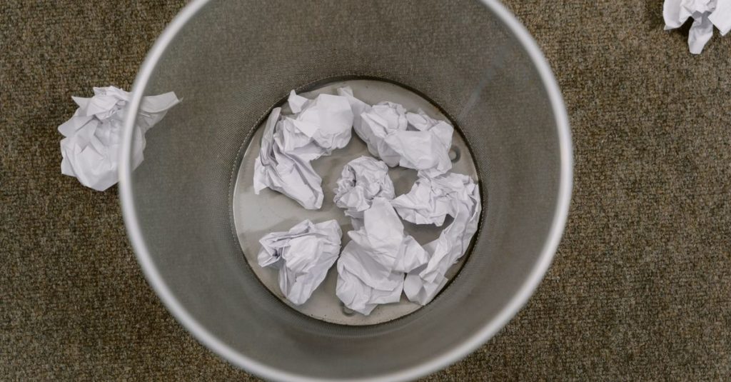 Overhead view of a trash can filled with crumpled paper on a carpeted floor.