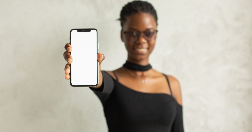 Positive African American female in black outfit and eyeglasses showing smartphone with blank screen while standing against white wall and looking at camera
