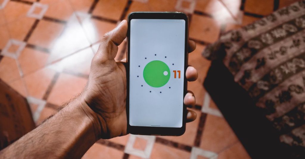 Close-up of a hand holding a smartphone displaying Android 11 interface indoors on patterned floor.