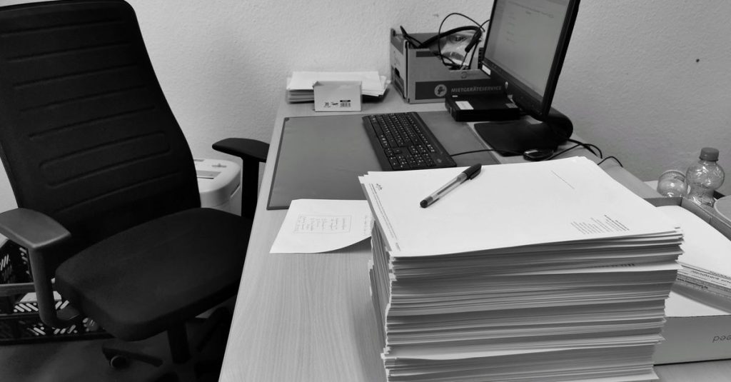 Black and white image of a cluttered office desk with a computer, chair, and paperwork.