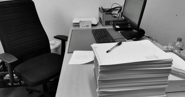 Black and white image of a cluttered office desk with a computer, chair, and paperwork.
