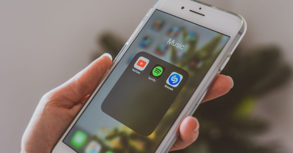 Hand holding a smartphone displaying popular music apps like Spotify and Shazam in a close-up shot.