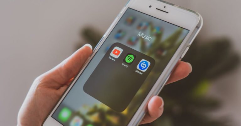 Hand holding a smartphone displaying popular music apps like Spotify and Shazam in a close-up shot.