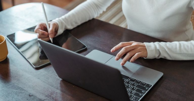 From above of crop anonymous businesswoman in white sweater working at table using tablet with stylus and typing on laptop