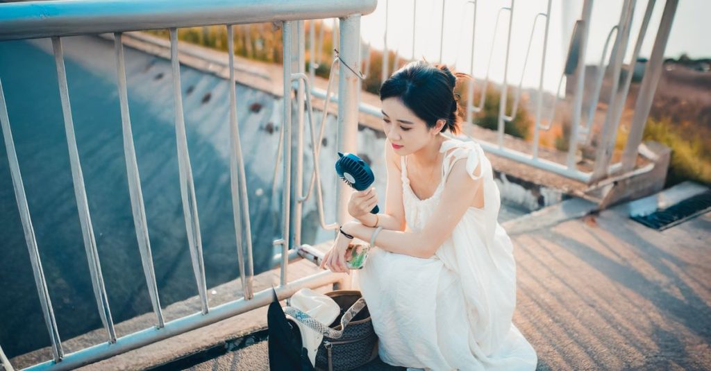 Woman in a white dress cools down with a portable fan on a sunny day outdoors.