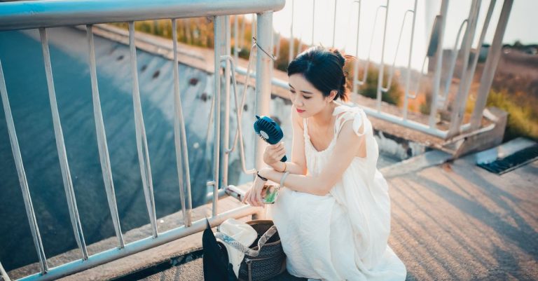 Woman in a white dress cools down with a portable fan on a sunny day outdoors.