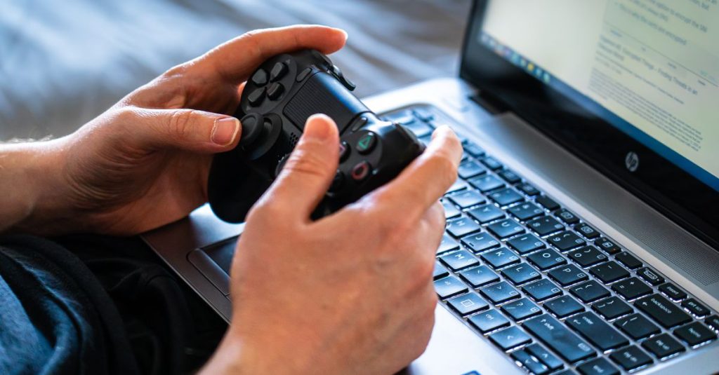 Person holds a gaming controller while interacting with a laptop, emphasizing modern gaming technology.
