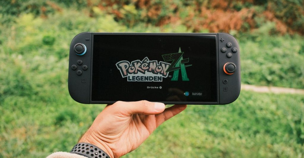 A hand holding a handheld gaming console displaying the Pokémon Legends game screen outdoors.