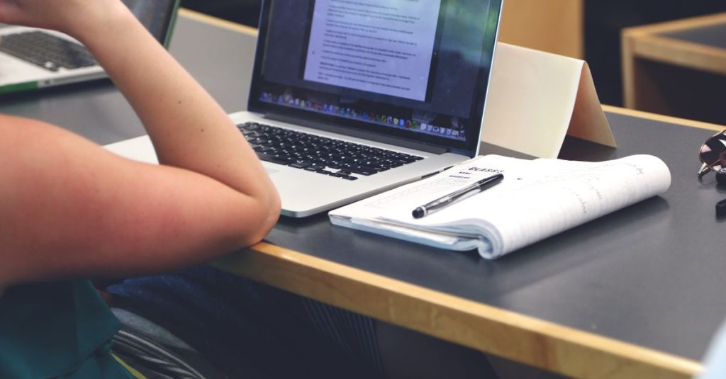 A student in a classroom setting using a laptop and taking notes in a notebook.