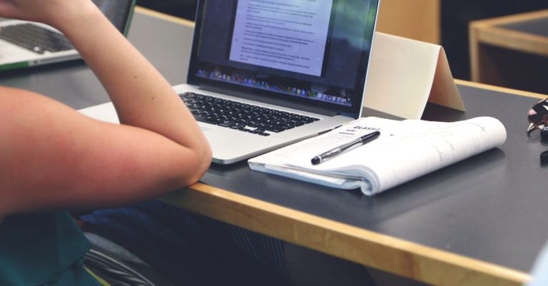 A student in a classroom setting using a laptop and taking notes in a notebook.