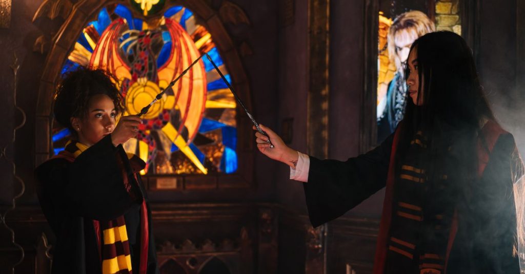 Two young women dressed in wizarding robes duel with wands in a mystical setting.