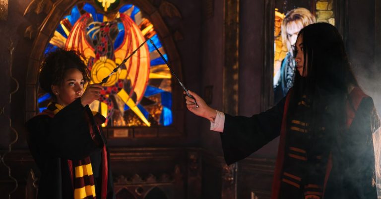 Two young women dressed in wizarding robes duel with wands in a mystical setting.