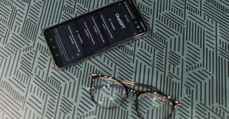 Close-up of smartphone displaying ChatGPT alongside eyeglasses on a patterned surface highlighting technology and style.