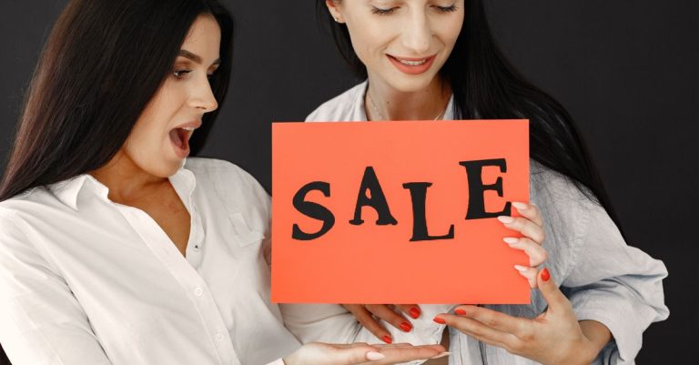 Two women energetically holding a 'SALE' sign, showcasing excitement and enthusiasm for a shopping event.