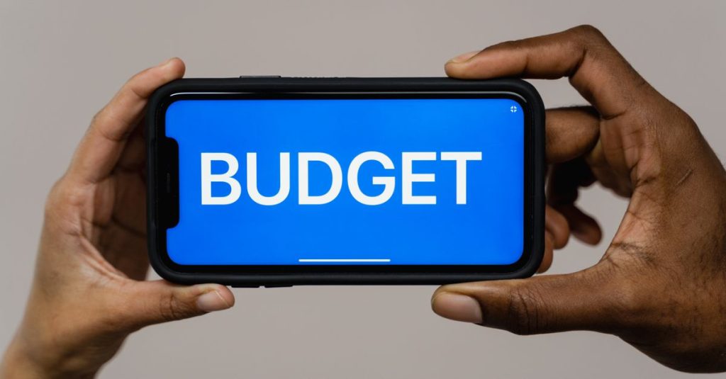 Two hands hold a smartphone displaying the word 'budget' on a blue screen, symbolizing financial planning.