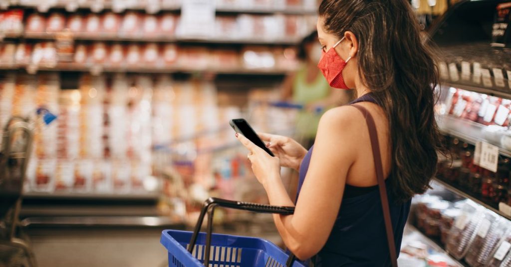 A woman with a mask shops in a grocery store using her smartphone and basket in hand.