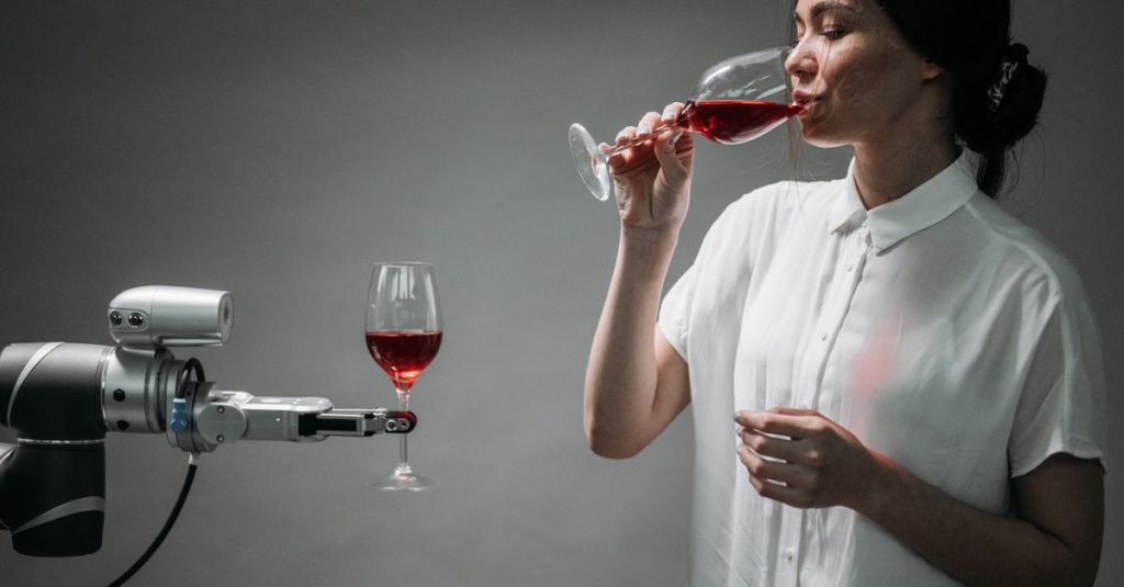 A woman in a white shirt drinking red wine interacts with a robot arm holding a wine glass.