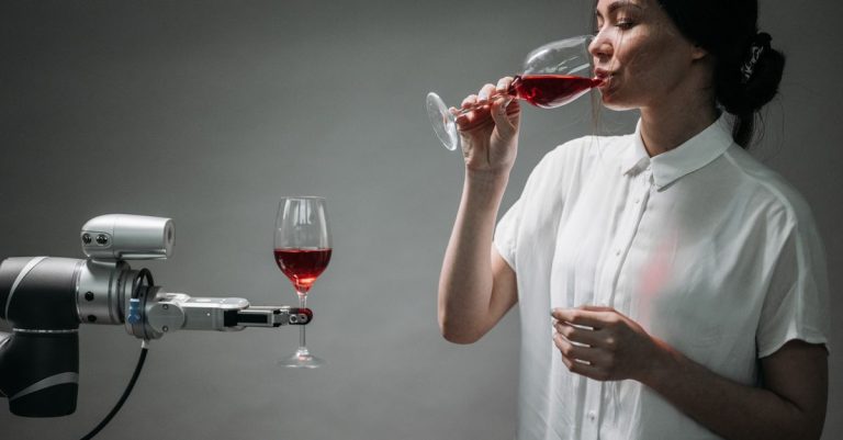 A woman in a white shirt drinking red wine interacts with a robot arm holding a wine glass.