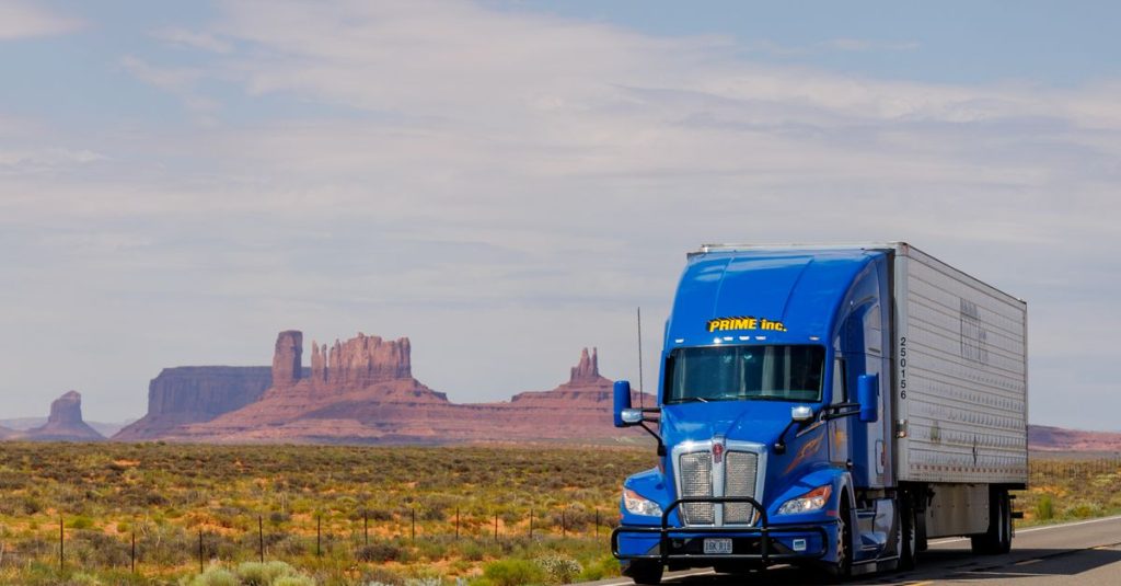 A blue truck travels on the highway with Monument Valley's iconic mesas in the background.