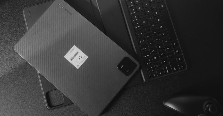 Monochrome image of a tablet, keyboard, and game controller on a black desk.