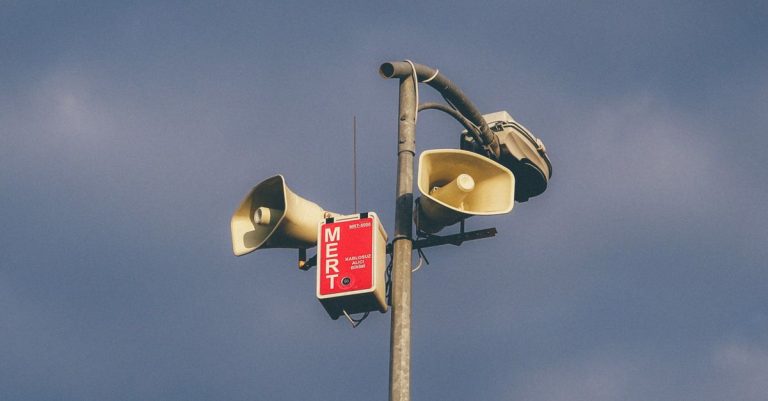 A warning system with loudspeakers on a pole against a cloudy sky, signaling emergency alerts.