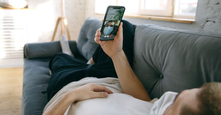 A man lying on a sofa using a smartphone for a video call while wearing a face mask indoors.