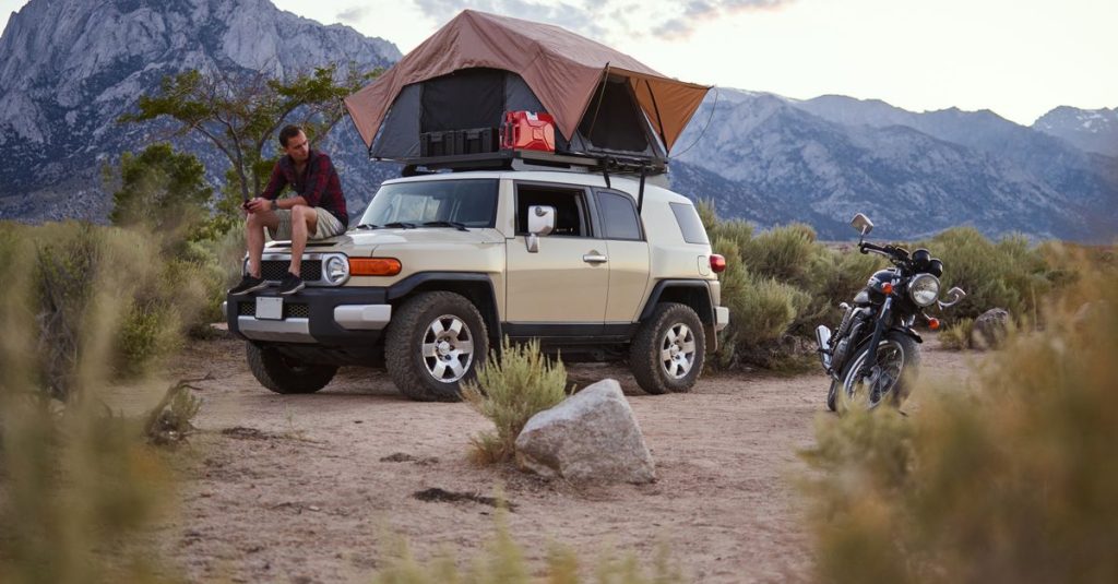 Outdoor camping scene with a man sitting on a car equipped with a rooftop tent, scenic mountain view.